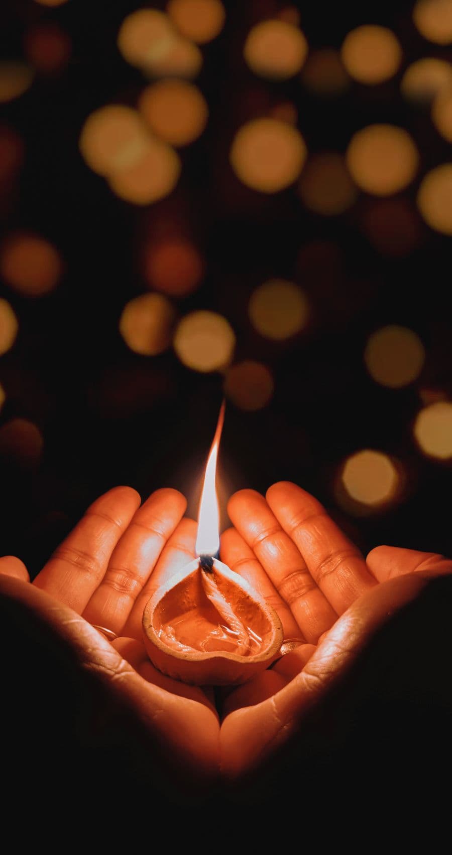 A lit diya cradled in cupped hands, warm lights glowing behind